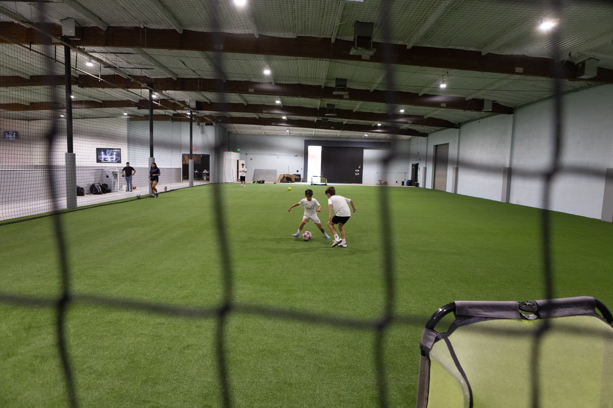 Two athletes train one-on-one on indoor turf, viewed through sideline netting, with staff and observers along the perimeter of the facility.