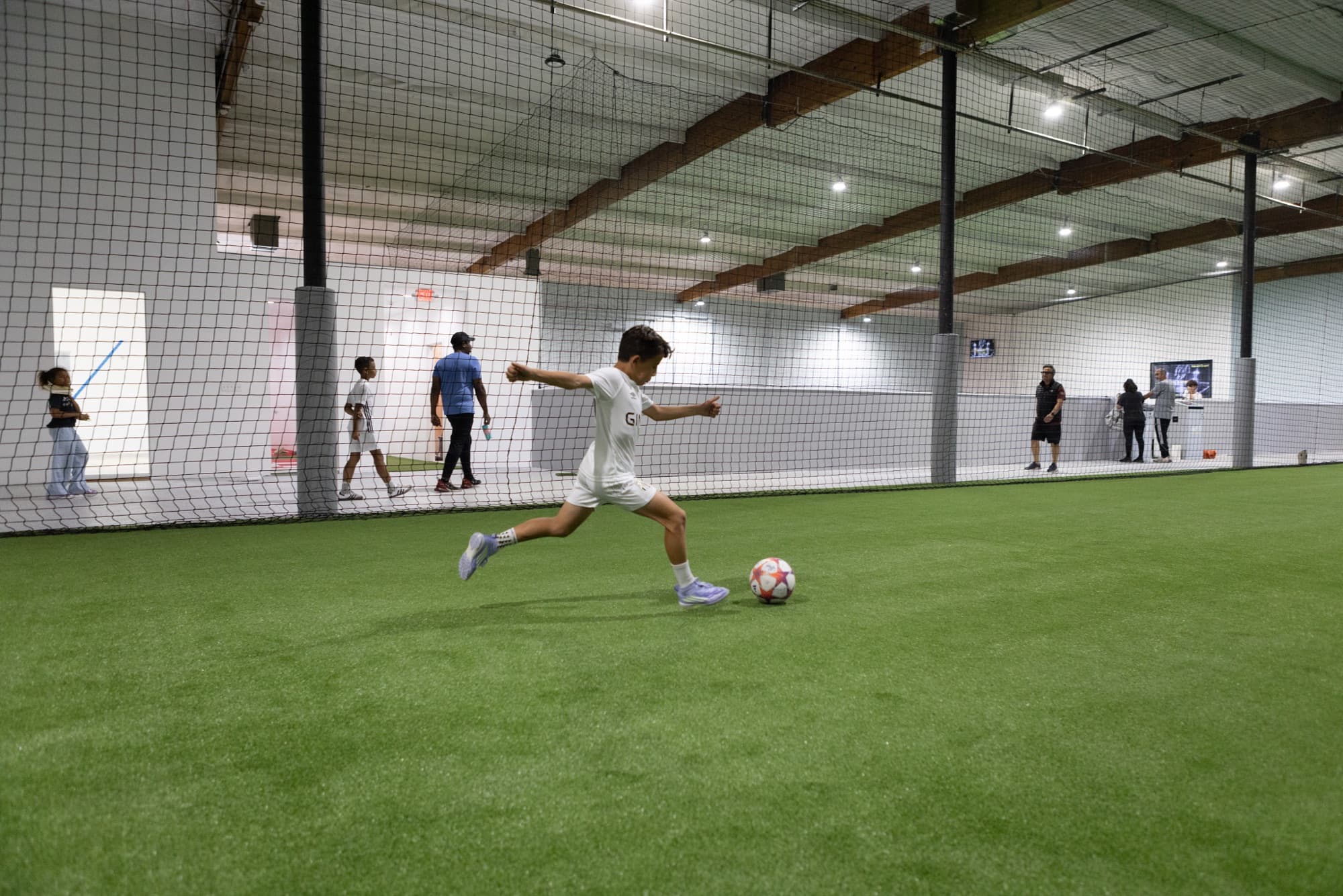 Athlete strikes a ball on an indoor turf field partitioned by netting, with additional training lanes visible in the background.